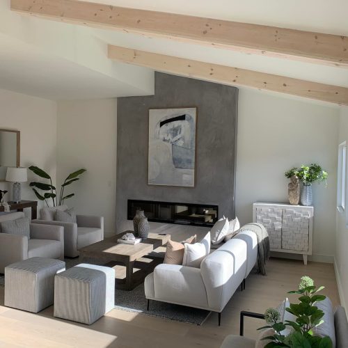 living room with white walls, foe finish fireplace accent wall, white ceiling with wood beams and grey couches.